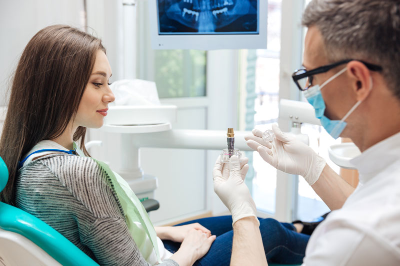 A dentist explains a dental implant model to a young woman in a dental chair, with an X-ray displayed behind them. She listens attentively, ready for treatment. A dentist explains a dental implant model to a young woman in a dental chair, with an X-ray displayed behind them. She listens attentively, ready for treatment.