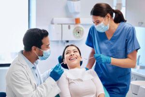 Happy dental patient smiling in the chair while a dentist and dental hygienist wearing masks and gloves perform a check-up in a modern dental clinic.